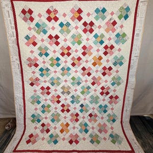 May include: A rectangular quilt featuring a repeating pattern of multi-colored fabric squares arranged in cross shapes. The quilt has a cream-colored background with a red border and an outer border printed with vintage-style text and graphics.