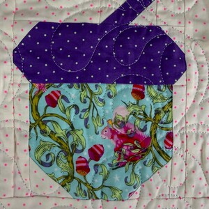 Puede incluir: Un dise&ntilde;o de bellota acolchada con una parte superior morada con lunares y una base azul claro con un patr&oacute;n de ardilla y flores. El fondo es blanco con lunares rosas y costuras decorativas.