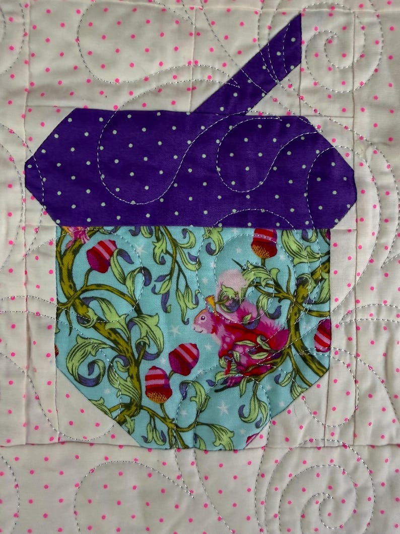 Puede incluir: Un dise&ntilde;o de bellota acolchada con una parte superior morada con lunares y una base azul claro con un patr&oacute;n de ardilla y flores. El fondo es blanco con lunares rosas y costuras decorativas.