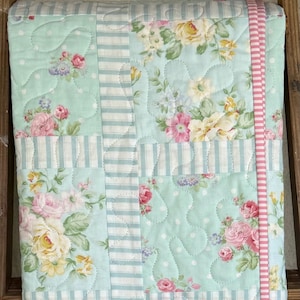 May include: A floral quilt featuring pink and yellow roses on a light blue background. The quilt has white polka dots and is bordered with light blue and white stripes and a pink ribbon.