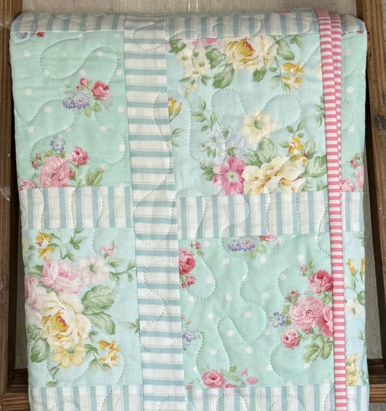 May include: A floral quilt featuring pink and yellow roses on a light blue background. The quilt has white polka dots and is bordered with light blue and white stripes and a pink ribbon.