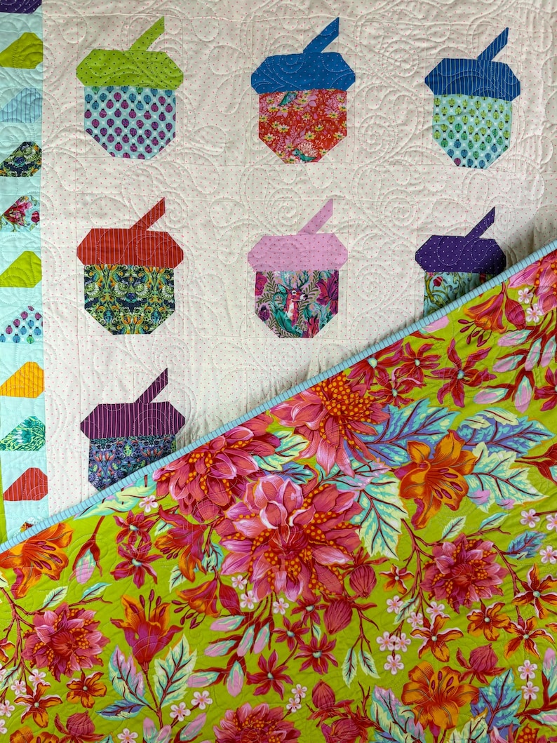 Puede incluir: Un edred&oacute;n colorido con formas de bellotas aplicadas en varias telas estampadas. El edred&oacute;n tiene un fondo blanco con un borde floral y un reverso floral verde vibrante. Las bellotas son en tonos de azul, verde, rojo y morado.