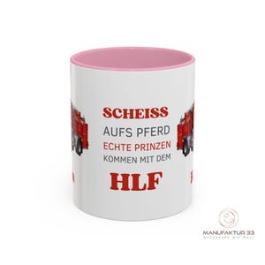 May include: White ceramic mug with a pink interior and the text "SCHEISS AUFS PFERD ECHTE PRINZEN KOMMEN MIT DEM HLF" in red. The mug features a red fire truck graphic on each side. The bottom right corner has a logo.