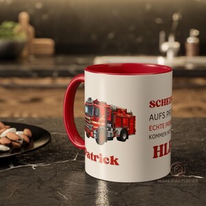 May include: A white ceramic mug with a red interior and handle. The mug features a red fire truck graphic and the name "Patrick" in red. Text in German is also visible on the mug.