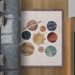 Può includere: Una coperta color crema con un design del sistema solare. I pianeti sono raffigurati come cerchi ad acquerello nei toni del blu, rosso, marrone e verde, con un sole giallo al centro. La coperta ha un bordo con frange.