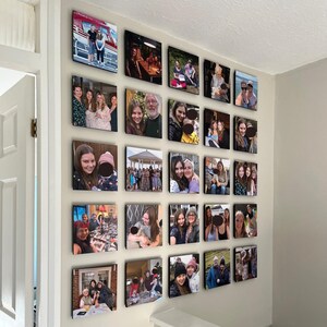 May include: A white wall with a gallery of framed photographs of different people and groups of people. The photographs are arranged in a grid pattern and are all in colour.