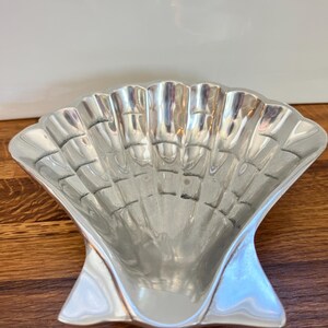 May include: A polished silver-toned metal dish shaped like a scallop shell. The bowl features a series of vertical ridges radiating from the base to the scalloped edge, creating a concave, fan-like form with a smooth, reflective metallic surface.