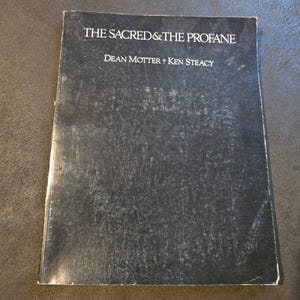 May include: A rectangular softcover book with a black, textured cover. The title THE SACRED & THE PROFANE is printed in white capital letters at the top, followed by the author names Dean Motter and Ken Steacy in white text below the title.