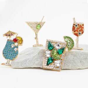 Könnte beinhalten: Vier goldfarbene Cocktail-Anstecker mit bunten Strasssteinen. Die Anstecker sind in Form eines Martini-Glases, eines Margarita-Glases, eines Weinglases und eines tropischen Getränks gestaltet.