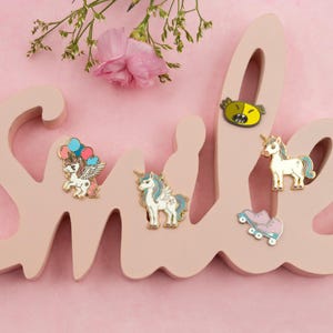 Könnte beinhalten: Fünf Emaille-Pins mit einem Einhorn mit Ballons, einem Einhorn mit einer Regenbogenmähne, einem Einhorn mit einer weißen Mähne, einem gelben Monstergesicht und einem Paar rosa Rollschuhen. Die Pins sind auf einem rosafarbenen Holzschild mit der Aufschrift "smile" angebracht.