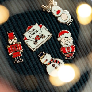 Puede incluir: Un conjunto de seis pines de esmalte con temas navideños. Los pines incluyen un reno, un muñeco de nieve, una carta a Santa Claus, un cascanueces y dos Papá Noeles. Los pines son todos de color dorado con detalles rojos, blancos y azules.
