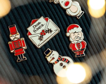 May include: A set of six enamel pins featuring Christmas themes. The pins include a reindeer, a snowman, a letter to Santa, a nutcracker, and two Santas. The pins are all gold-toned with red, white, and blue accents.