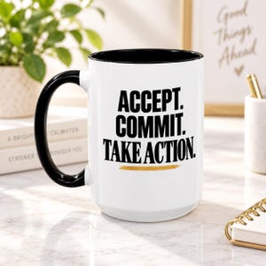 May include: A white ceramic mug with a black handle and interior rim. The front features bold black text that reads ACCEPT. COMMIT. TAKE ACTION. Below the text is a horizontal gold-colored brushstroke graphic. The mug has a cylindrical shape.