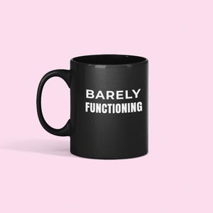 May include: A black ceramic mug with a curved handle. The mug features the white text "BARELY FUNCTIONING" in a bold, sans-serif font. The mug is set against a light pink background.
