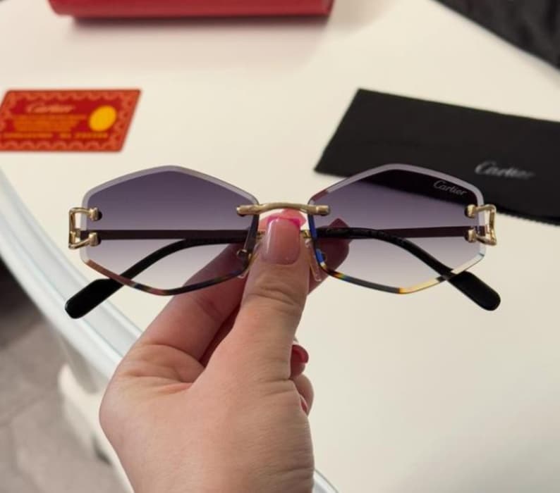 May include: Gold-rimmed sunglasses with a dark gray gradient lens. The sunglasses have a geometric shape and the word 'Cartier' is printed on the right lens.