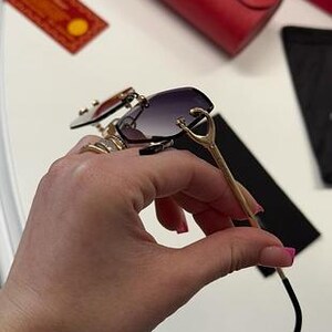May include: Gold-tone sunglasses with a black frame and dark gray lenses. The sunglasses have a unique design with a curved frame and a decorative detail on the temples.