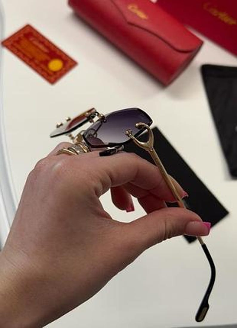 May include: Gold-tone sunglasses with a black frame and dark gray lenses. The sunglasses have a unique design with a curved frame and a decorative detail on the temples.