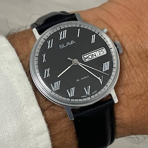 Puede incluir: Un reloj de pulsera Slava vintage con esfera negra, manecillas plateadas y números romanos. El reloj muestra el día y la fecha, con una correa de cuero negro. La esfera del reloj dice "SLAVA" y "26 JEWELS".