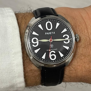 Puede incluir: Un reloj de pulsera Paketa con esfera negra, números y agujas blancas, y un segundero rojo. El reloj tiene una caja de acero inoxidable y una correa de cuero negro. La esfera incluye el texto "PAKETA" y texto cirílico.
