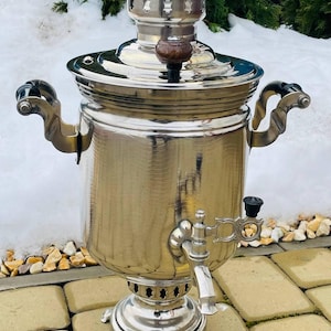 May include: A polished silver samovar, a traditional Russian tea urn, with a decorative tap and handles. It features a round base and a lid with a wooden knob. The samovar is designed for making tea.