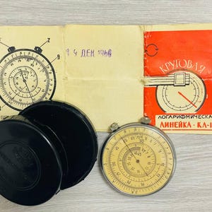 May include: A vintage circular slide rule with a silver frame and cream dial, alongside a black case and paperwork. The paperwork is dated "19 ДЕК 1966" and features Russian text. The slide rule has various scales for calculations.