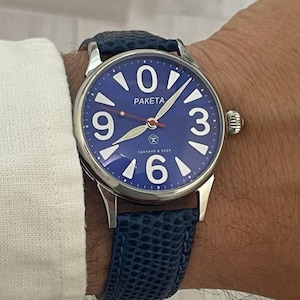 Puede incluir: Un reloj de pulsera con esfera azul y correa de cuero azul. La esfera del reloj tiene números y agujas blancas, con la marca "PAKETA". El reloj está en una muñeca, con una manga de camisa blanca visible.