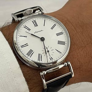 Puede incluir: Un reloj de bolsillo plateado con esfera blanca y números romanos negros. El reloj tiene un pequeño dial de segundos y una correa de cuero negro. La esfera del reloj dice "Molnija".