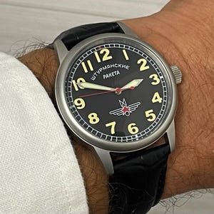 Puede incluir: Reloj de pulsera redondo con esfera negra, números y agujas doradas. El reloj tiene una caja de acero inoxidable y una correa de cuero negro. La esfera del reloj incluye el texto "Штурманские РАКЕТА" y un emblema rojo y blanco.
