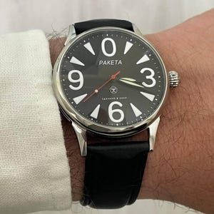 Puede incluir: Un reloj de pulsera plateado con esfera negra y correa de cuero negro. La esfera muestra números blancos y la palabra "PAKETA". El reloj está en una muñeca.