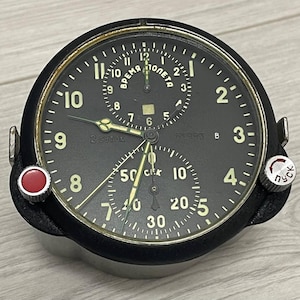 Könnte beinhalten: Eine schwarze Vintage-Analog-Uhr mit weißen Ziffern und Zeigern. Das Zifferblatt hat mehrere Zifferblätter und den Text "ВРЕМЯ ПОЛЕТА" und "2 суток". Auf der linken Seite befindet sich ein roter Knopf und auf der rechten Seite ein silberner Knopf mit kyrillischem Text.