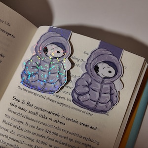 May include: Two Snoopy-themed bookmarks featuring Snoopy in a puffy winter coat. The bookmarks are purple and white, with a glittery effect on the coat. The bookmarks are placed on the pages of a book.