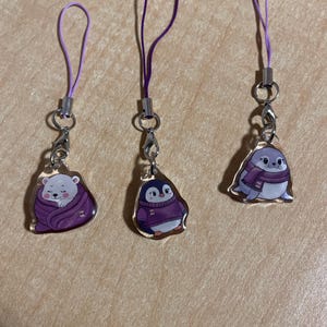 May include: Three acrylic charms with purple lanyards. The charms depict a polar bear wrapped in a purple blanket, a penguin in a purple sweater, and a seal wearing a purple scarf. Each charm has a silver clasp.