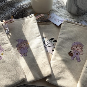 May include: Several beige canvas zipper pouches, each embroidered with a different whimsical design. Designs include a purple airplane, a witch, a character with a dog, a girl with a basket, and a bunny with a carrot.
