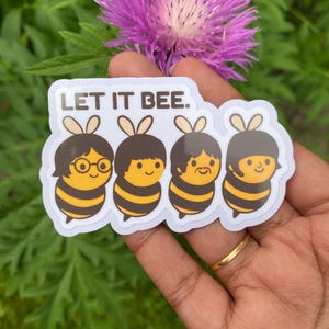 Puede incluir: Una pegatina blanca con el texto "LET IT BEE." presenta cuatro abejas de dibujos animados con cabello castaño y cuerpos a rayas amarillas y negras. La pegatina se sostiene en una mano con un anillo dorado, con una flor morada y follaje verde en el fondo.