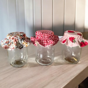 Op de afbeelding: Drie glazen potten met stoffen deksels. De deksels zijn in verschillende patronen: bloemen, rood-wit geruit en roze met aardbeien.