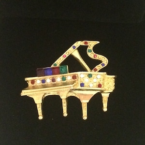 May include: A gold-tone brooch shaped like a grand piano with colourful rhinestones. The piano has a black and white keyboard, and the rhinestones are red, blue, green, and clear.