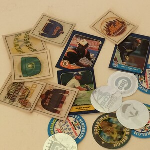 Könnte beinhalten: Eine Sammlung von Baseball-Sammelkarten und Aufklebern mit verschiedenen Teams und Spielern, darunter die Mets, Brewers und Cleveland Indians. Die Karten sind in verschiedenen Farben und zeigen Bilder von Spielern in Aktion. Es gibt auch mehrere runde Aufkleber mit Teamlogos und Spielernamen.