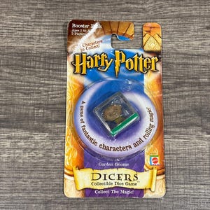 May include: A Harry Potter themed collectible dice game featuring a Garden Gnome character. The dice is in a clear plastic container with a green base. The packaging is yellow and orange with the words "Harry Potter" and "Dicers" printed on it.