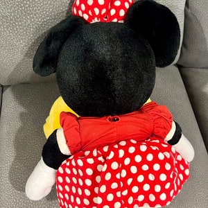 K&ouml;nnte beinhalten: Ein Pl&uuml;schtier von Minnie Mouse, das ein rotes Kleid mit wei&szlig;en Punkten und eine rote Schleife mit wei&szlig;en Punkten tr&auml;gt. Das Spielzeug sitzt auf einer grauen Oberfl&auml;che.