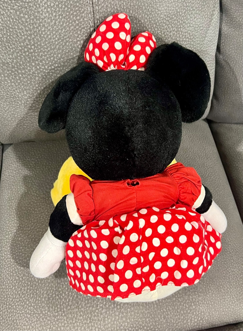 K&ouml;nnte beinhalten: Ein Pl&uuml;schtier von Minnie Mouse, das ein rotes Kleid mit wei&szlig;en Punkten und eine rote Schleife mit wei&szlig;en Punkten tr&auml;gt. Das Spielzeug sitzt auf einer grauen Oberfl&auml;che.