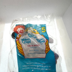 Puede incluir: Una bolsa de pl&aacute;stico que contiene un juguete de peluche de Minnie Mouse. El juguete es morado y negro con un lazo blanco. La bolsa es azul y amarilla con las palabras "Mickey Mania" y "Minnie Mouse Juguete Suave" impresas en ella.