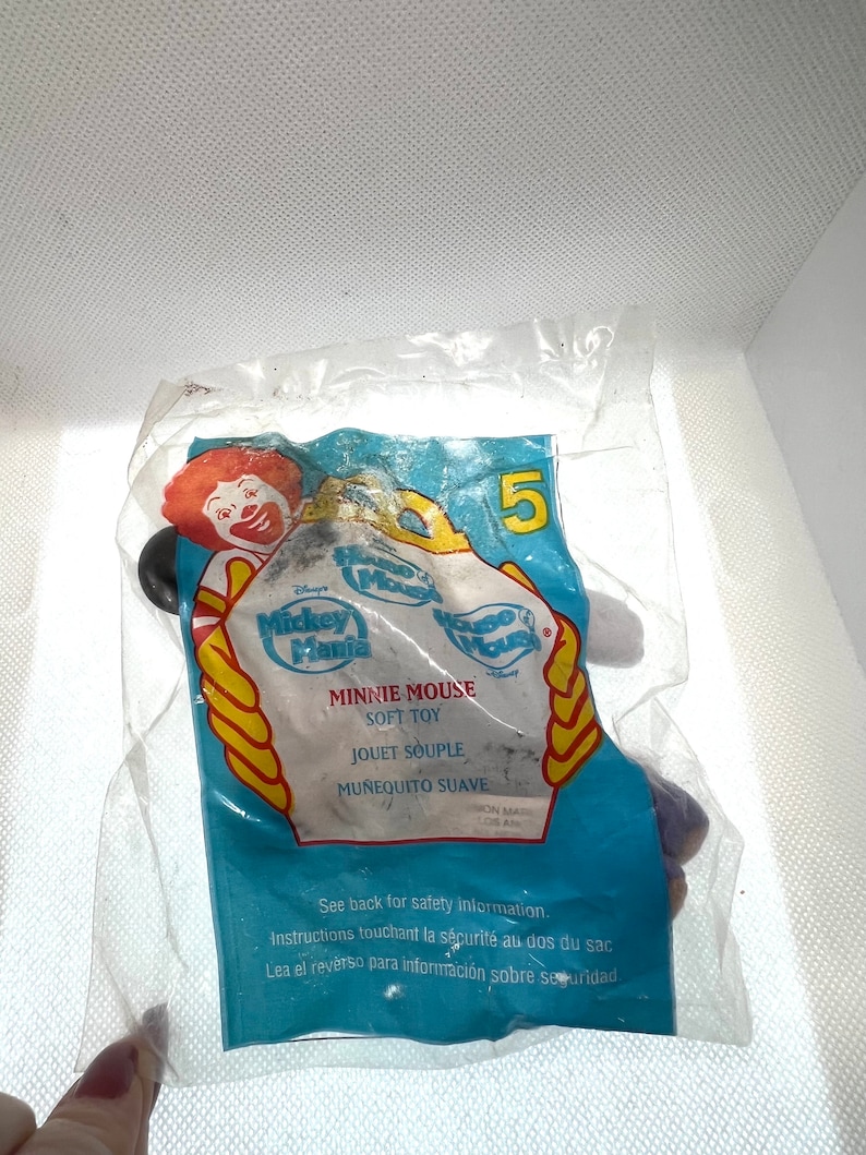 Puede incluir: Una bolsa de pl&aacute;stico que contiene un juguete de peluche de Minnie Mouse. El juguete es morado y negro con un lazo blanco. La bolsa es azul y amarilla con las palabras "Mickey Mania" y "Minnie Mouse Juguete Suave" impresas en ella.