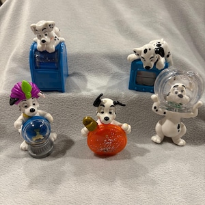 May include: Five Dalmatian dog figurines, each with a different pose and accessory. The figurines are made of plastic and are painted in black and white. One figurine is holding a blue mailbox, another is holding a red and white ornament, another is holding a snow globe, another is sitting on a blue mailbox, and another is sitting on a blue and white box.