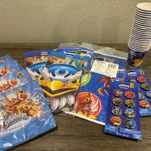 May include: A selection of Skylanders party supplies, including napkins, plates, cups, and button badges. The items feature vibrant character designs in blue, red, and yellow. The cups are white with a blue character design.