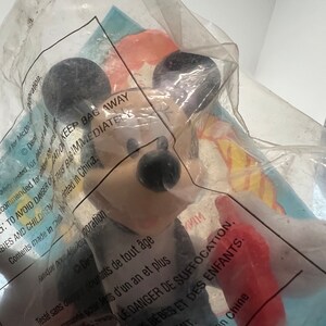 Puede incluir: Una figura de Mickey Mouse roja y negra con una camisa a rayas blancas y negras. La figura est&aacute; empaquetada en una bolsa de pl&aacute;stico transparente con una etiqueta de advertencia que dice "KEEP BAG AWAY TO AVOID DANGER OF SUFFOCATION.  MISE EN GARDE LE DANGER DE SUFFOCATION.  Contents made in China. Printed in China."