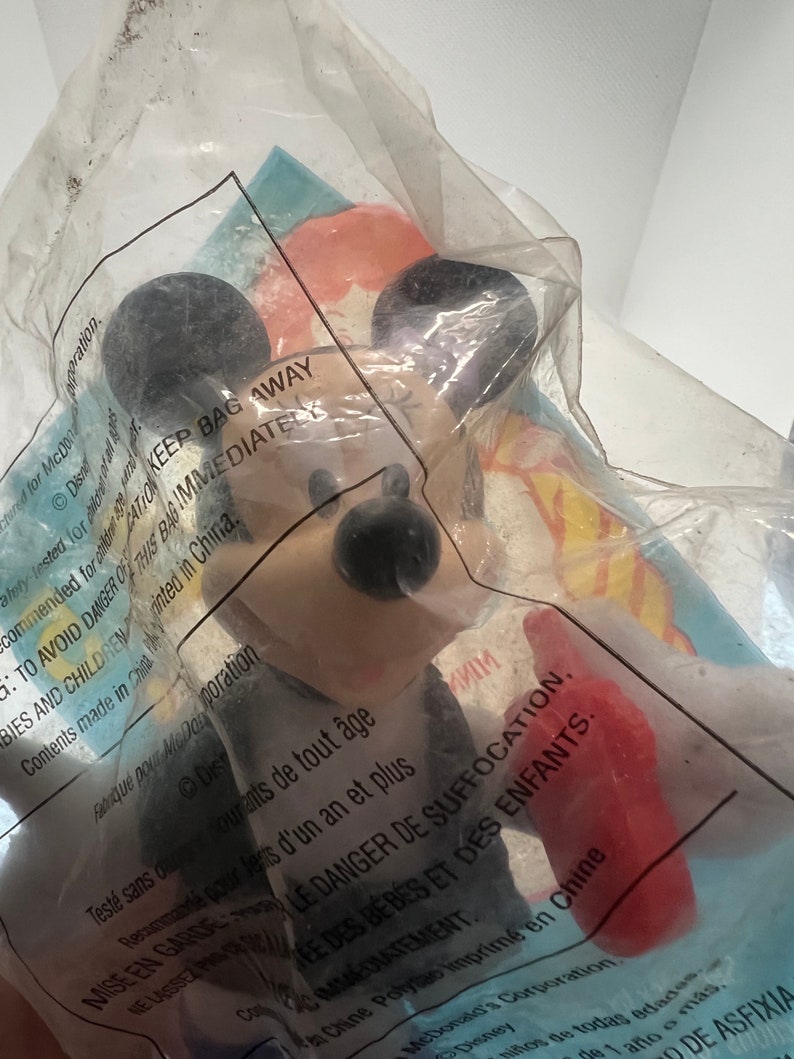 Puede incluir: Una figura de Mickey Mouse roja y negra con una camisa a rayas blancas y negras. La figura est&aacute; empaquetada en una bolsa de pl&aacute;stico transparente con una etiqueta de advertencia que dice "KEEP BAG AWAY TO AVOID DANGER OF SUFFOCATION.  MISE EN GARDE LE DANGER DE SUFFOCATION.  Contents made in China. Printed in China."