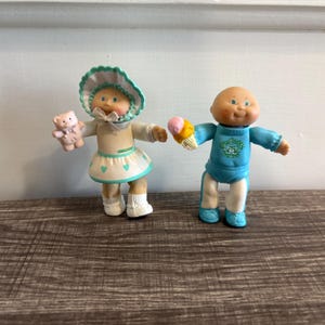 Puede incluir: Dos muñecas vintage de Cabbage Patch Kids. Una muñeca viste un vestido blanco con una falda turquesa con estampado de corazones y un gorro a juego, sosteniendo un osito de peluche rosa. La otra muñeca lleva un conjunto azul y sostiene un cono de helado.