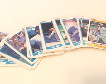 May include: A collection of baseball trading cards featuring various players from the 1980s. The cards are arranged in a fan shape, with the top card showing a player in a blue uniform with the text "10th Anniversary George Bell" on it.