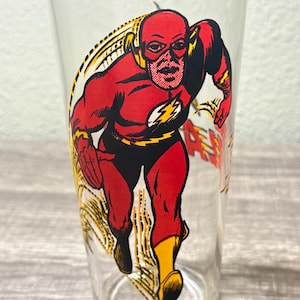 May include: Clear glass featuring a vintage illustration of The Flash in a running pose. The superhero is depicted in a red and black costume with a yellow lightning bolt emblem. The glass has a classic design.