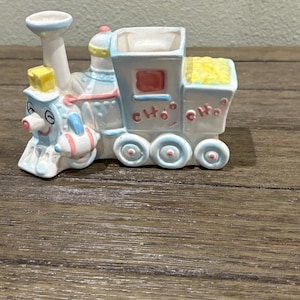 May include: A ceramic train figurine in pastel colors. The train features a smiling face, a yellow square, and the word "Cho" on the side. The wheels and other details are painted in blue, pink, and white.